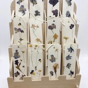May include: A wooden display stand holds several paper bookmarks, each decorated with pressed flowers in various colours. The flowers include blue, purple, and yellow blooms, and are attached with a cream-coloured string. The stand is made of light-coloured wood.