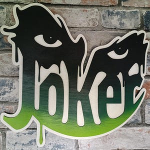 May include: Wooden sign spelling "Joker" in a dripping font, with a black and green gradient design. The sign has a white outline and is mounted on a brick wall.
