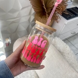 May include: A clear glass reusable can with a bamboo lid and pink lettering that reads "BIG BUTT ENERGY". The can is filled with a brown liquid and has a pink straw sticking out of the lid.