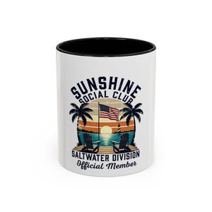 May include: White ceramic mug with a black interior. The mug features the text "SUNSHINE SOCIAL CLUB" above a beach scene with palm trees, two chairs, and an American flag. The text "SALTWATER DIVISION Official Member" is below the scene.