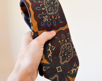 May include: A patterned tie featuring a paisley design in shades of gold, navy, burgundy, and brown. The tie is held up against a neutral background, showcasing the intricate floral pattern. This classic accessory is suitable for formal or business attire.