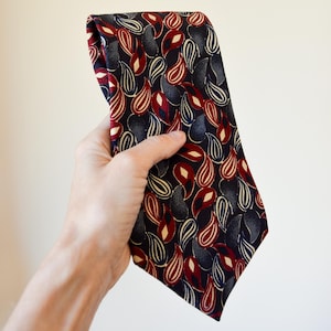 May include: A folded necktie with a dark navy background and a repeating paisley pattern in red, white, and gray. The tie is held up against a plain, off-white background.
