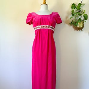 May include: A vibrant fuchsia velvet gown with short sleeves and a scoop neckline. The dress features a white band with gold and copper-colored embellishments at the waist. The floor-length dress is displayed on a mannequin.