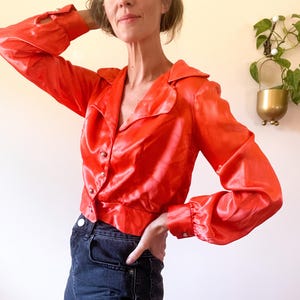 May include: A vibrant, red satin blouse with long sleeves and a collared neckline. The blouse features button closures and is paired with dark denim shorts. The outfit is a bold fashion statement.