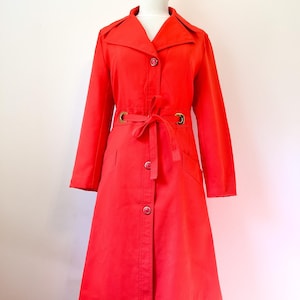 Peut inclure: Un trench-coat rouge vif avec une fermeture boutonnée, une ceinture assortie et des boucles de ceinture dorées. Le manteau est présenté sur un mannequin en bois. Il a des manches longues et un col classique.