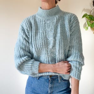 May include: A light blue cable-knit sweater with a mock neck. The sweater has a textured appearance and is paired with blue jeans. The sleeves are long and the overall style is casual and comfortable.