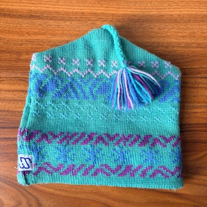 May include: A turquoise knitted hat featuring a geometric pattern in purple and blue. The hat has a tassel with blue, purple, and white strands. A small white tag with a blue logo is sewn on the side.