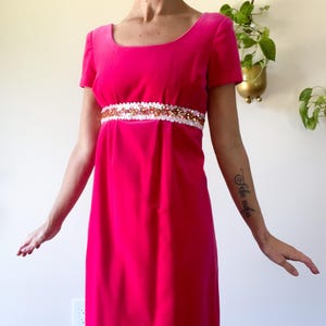 May include: A vibrant fuchsia-colored dress with short sleeves and a scoop neckline. The dress features a decorative white and gold embellished band at the waist. The dress is a vintage style, likely from the 1960s or 1970s.