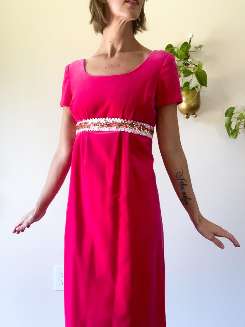May include: A vibrant fuchsia-colored dress with short sleeves and a scoop neckline. The dress features a decorative white and gold embellished band at the waist. The dress is a vintage style, likely from the 1960s or 1970s.