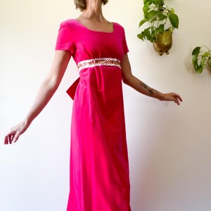 May include: A vibrant pink velvet gown with short sleeves and a square neckline. The dress features a decorative embellished belt with white and gold accents. A large bow detail is visible at the back.