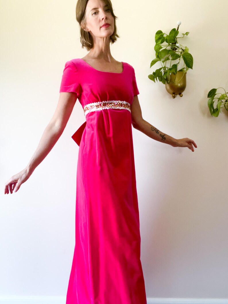 May include: A vibrant pink velvet gown with short sleeves and a square neckline. The dress features a decorative embellished belt with white and gold accents. A large bow detail is visible at the back.