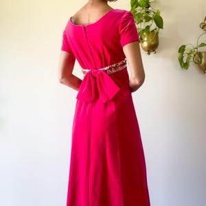 May include: A vibrant fuchsia velvet dress with short sleeves and a floor-length skirt. The dress features a decorative embellished belt and a large bow at the back. The dress has a scoop neckline and a zipper closure.