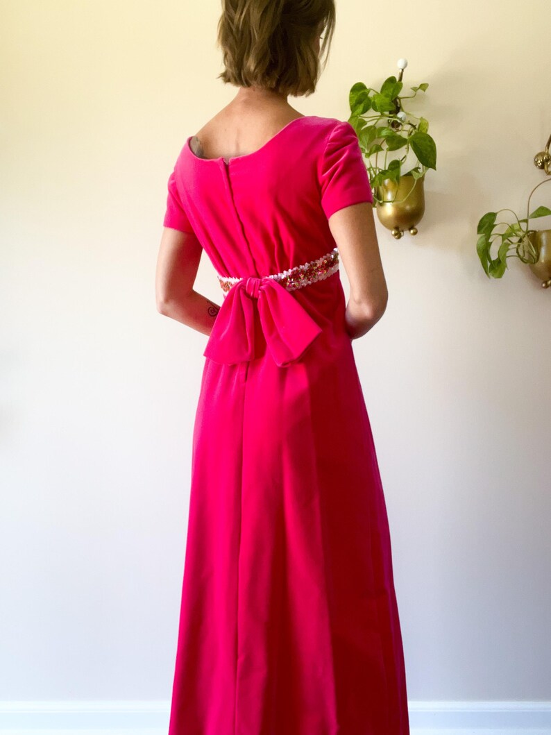 May include: A vibrant fuchsia velvet dress with short sleeves and a floor-length skirt. The dress features a decorative embellished belt and a large bow at the back. The dress has a scoop neckline and a zipper closure.