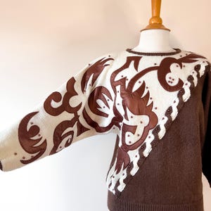 Può includere: Un maglione di lana marrone e bianco con un design in pelle marrone sulla manica sinistra e un bordo in pelle marrone sul lato destro. Il maglione ha un collo rotondo e maniche lunghe.
