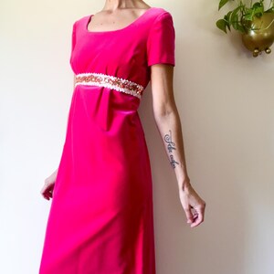 May include: A bright pink, floor-length dress with short sleeves and a square neckline. The dress features a white and sequined band at the waist. The dress is made of a soft, velvet-like material.