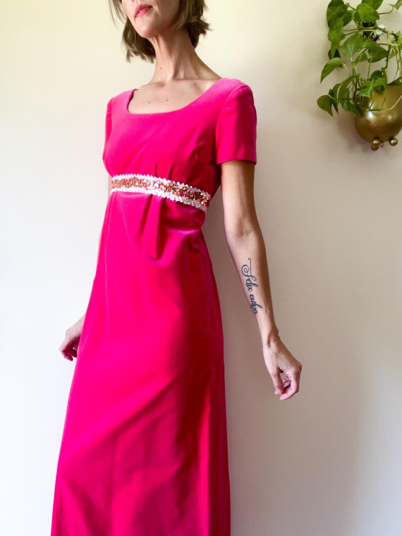 May include: A bright pink, floor-length dress with short sleeves and a square neckline. The dress features a white and sequined band at the waist. The dress is made of a soft, velvet-like material.