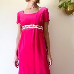May include: A vibrant fuchsia-colored dress with short sleeves and a square neckline. The dress features a white and gold embellished band at the waist. The long, flowing skirt adds to the vintage style of the garment.