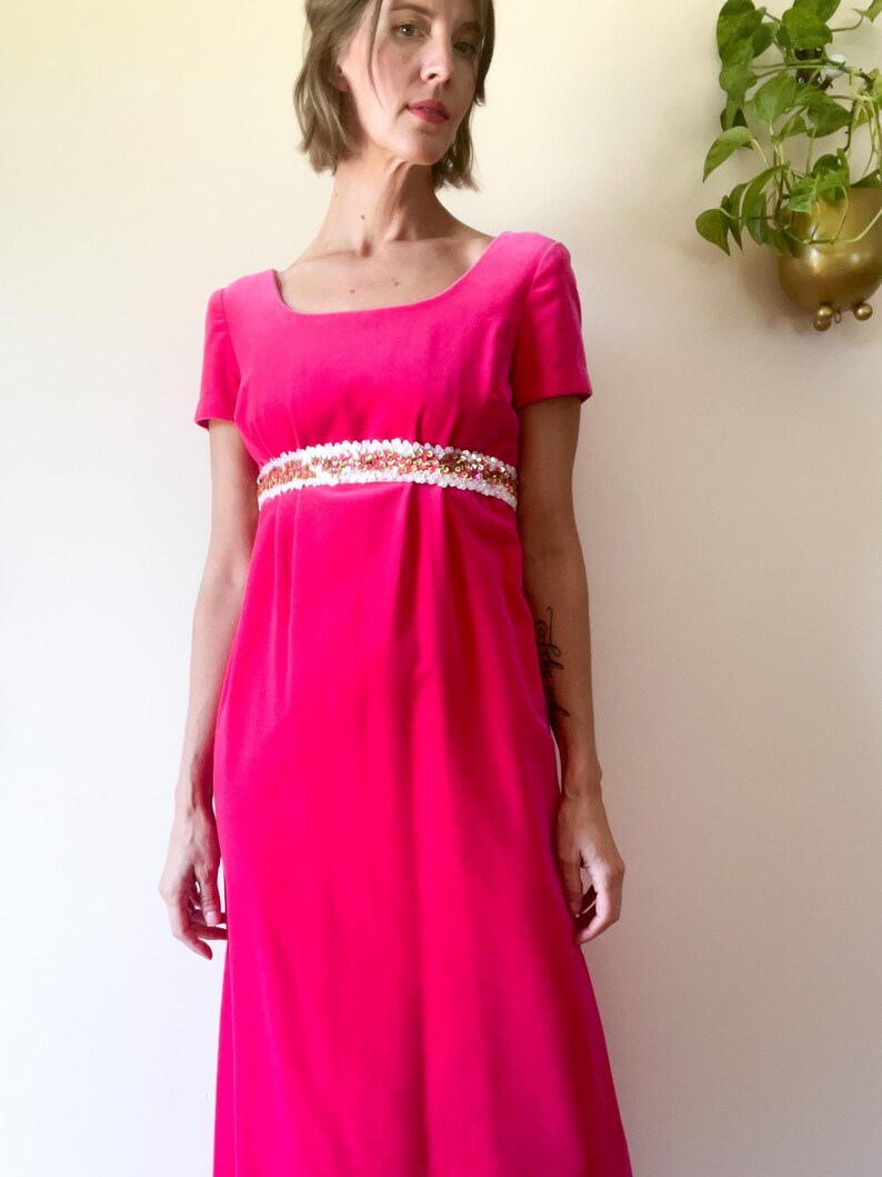 May include: A vibrant fuchsia-colored dress with short sleeves and a square neckline. The dress features a white and gold embellished band at the waist. The long, flowing skirt adds to the vintage style of the garment.