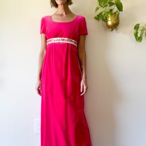 May include: A bright pink, floor-length dress with short sleeves and a square neckline. The dress features a decorative white and gold embellished band at the waist. The dress is made of a soft, velvet-like material.