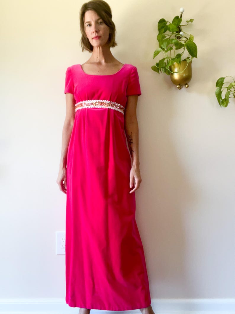 May include: A bright pink, floor-length dress with short sleeves and a square neckline. The dress features a decorative white and gold embellished band at the waist. The dress is made of a soft, velvet-like material.