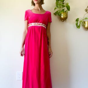 May include: A vibrant fuchsia-colored, floor-length dress with short sleeves. The dress features a scoop neckline and a decorative white and gold embellished band at the waist. The dress is made of a soft, flowing fabric.