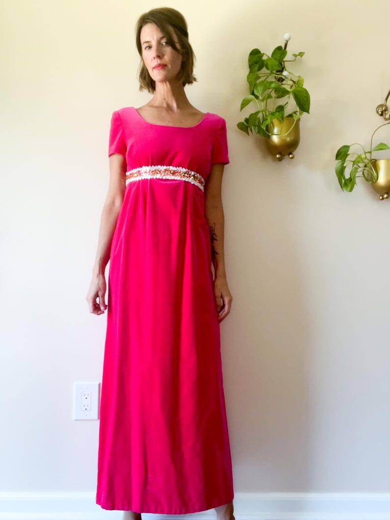 May include: A vibrant fuchsia-colored, floor-length dress with short sleeves. The dress features a scoop neckline and a decorative white and gold embellished band at the waist. The dress is made of a soft, flowing fabric.