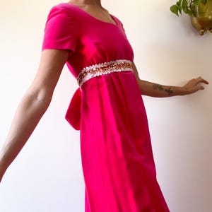 May include: A bright pink, short-sleeved dress with a square neckline and a decorative belt. The belt is white with gold and pink embellishments. The dress has a bow in the back.