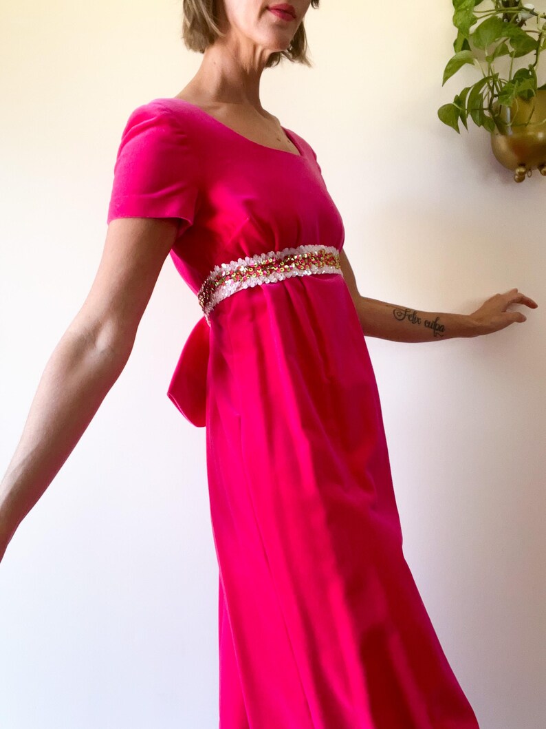 May include: A bright pink, short-sleeved dress with a square neckline and a decorative belt. The belt is white with gold and pink embellishments. The dress has a bow in the back.