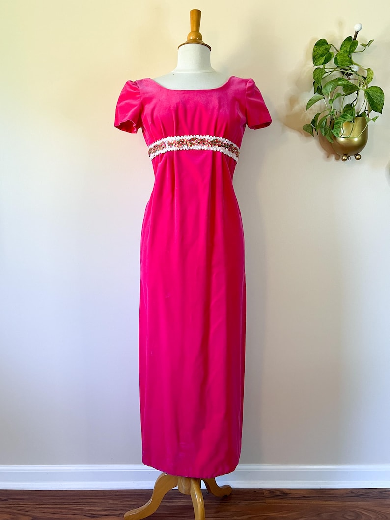 May include: A vibrant fuchsia velvet gown with short sleeves and a scoop neckline. The dress features a white band with gold and copper-colored embellishments at the waist. The floor-length dress is displayed on a mannequin.