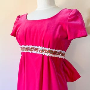 May include: A bright pink velvet dress with short sleeves and a scoop neckline. The dress features a white and sequined belt with red and green accents. The dress is displayed on a mannequin.