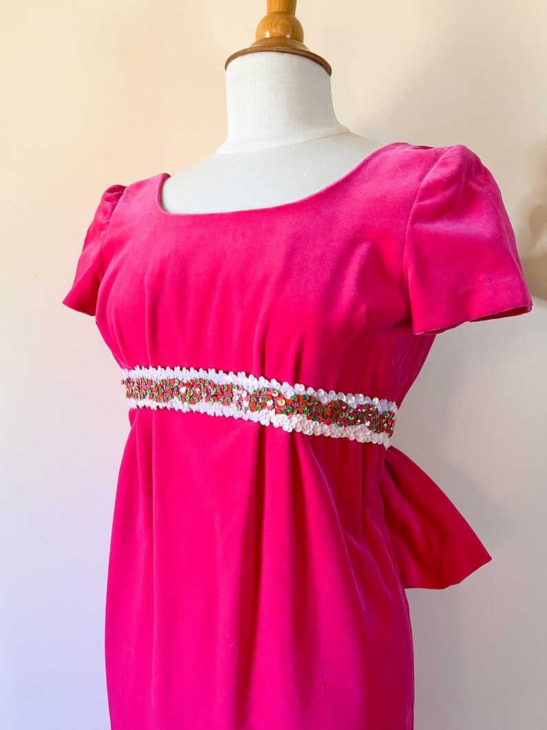 May include: A bright pink velvet dress with short sleeves and a scoop neckline. The dress features a white and sequined belt with red and green accents. The dress is displayed on a mannequin.