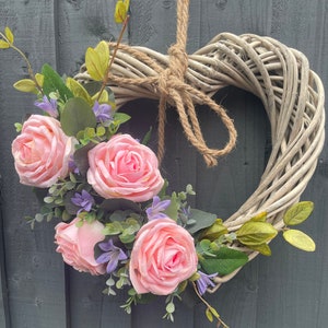 May include: A white wicker heart-shaped wreath decorated with pink roses and purple flowers.