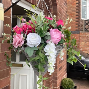 May include: A grey hanging basket filled with pink and white artificial flowers, including roses, peonies, and wisteria. The basket is hanging from a black metal hook attached to a brick wall.