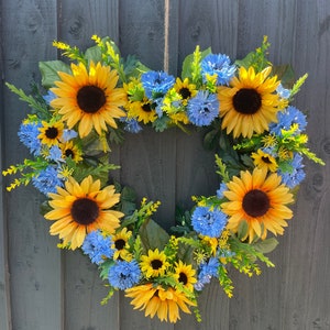 May include: A heart-shaped wreath made of sunflowers and blue flowers. The wreath is hanging on a grey wooden fence.