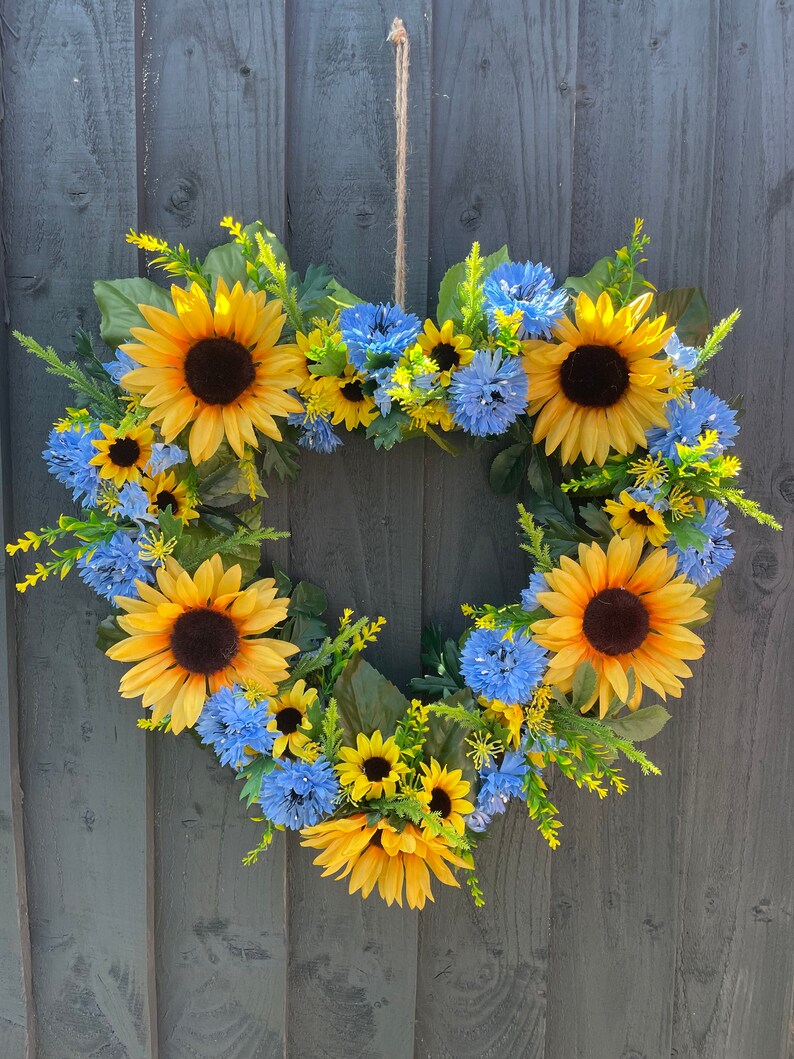 May include: A heart-shaped wreath made of sunflowers and blue flowers. The wreath is hanging on a grey wooden fence.