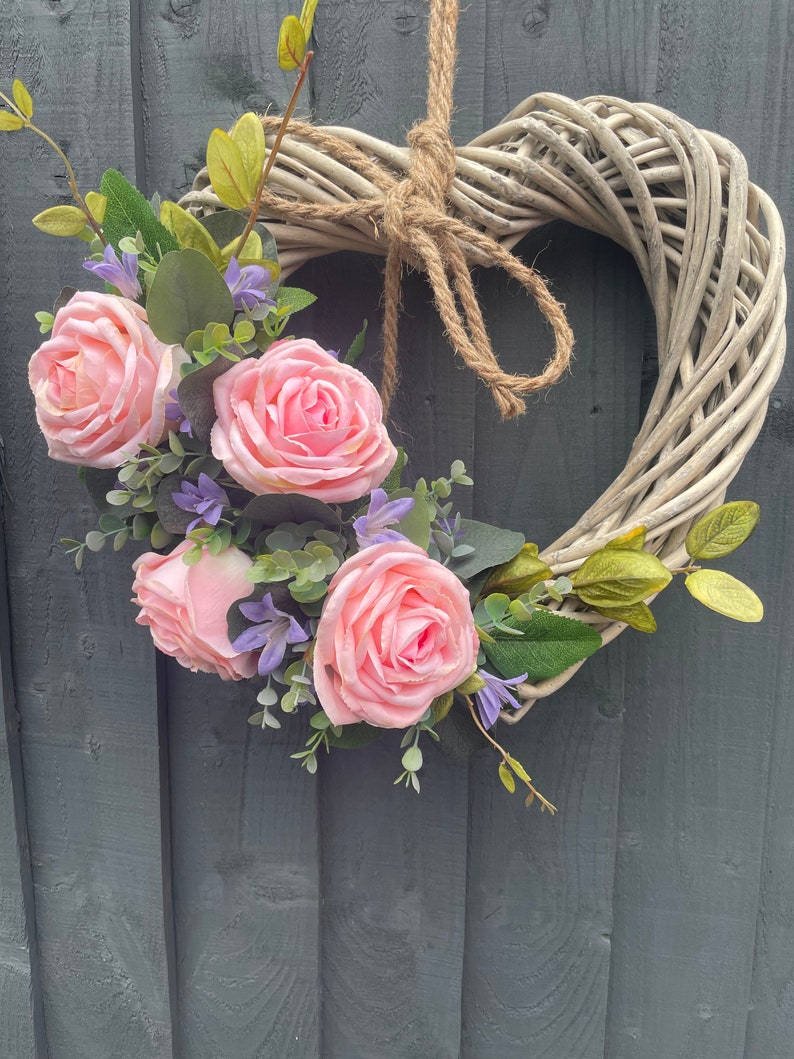 May include: A white wicker heart-shaped wreath decorated with pink roses and purple flowers.