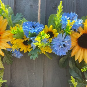May include: A wreath made of artificial sunflowers and blue flowers. The wreath is hanging on a grey wooden fence.
