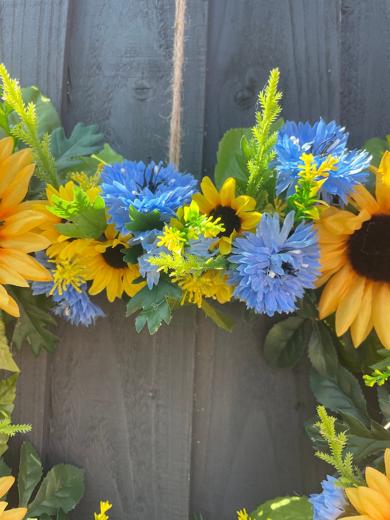 May include: A wreath made of artificial sunflowers and blue flowers. The wreath is hanging on a grey wooden fence.