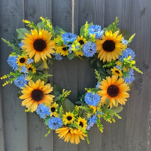 May include: A heart-shaped wreath made of sunflowers and blue flowers. The wreath is hanging on a wooden fence.