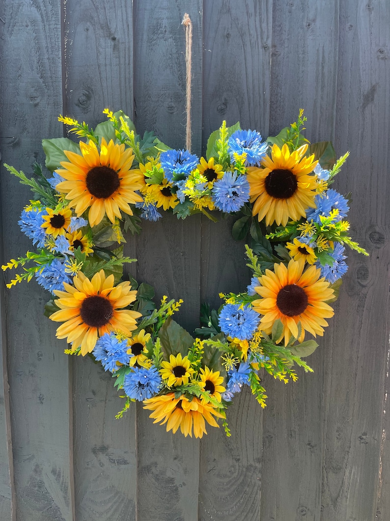 May include: A heart-shaped wreath made of sunflowers and blue flowers. The wreath is hanging on a wooden fence.