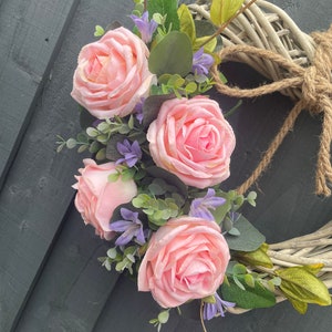May include: A heart-shaped wicker wreath decorated with pink roses and purple flowers. The wreath is tied with a brown twine.
