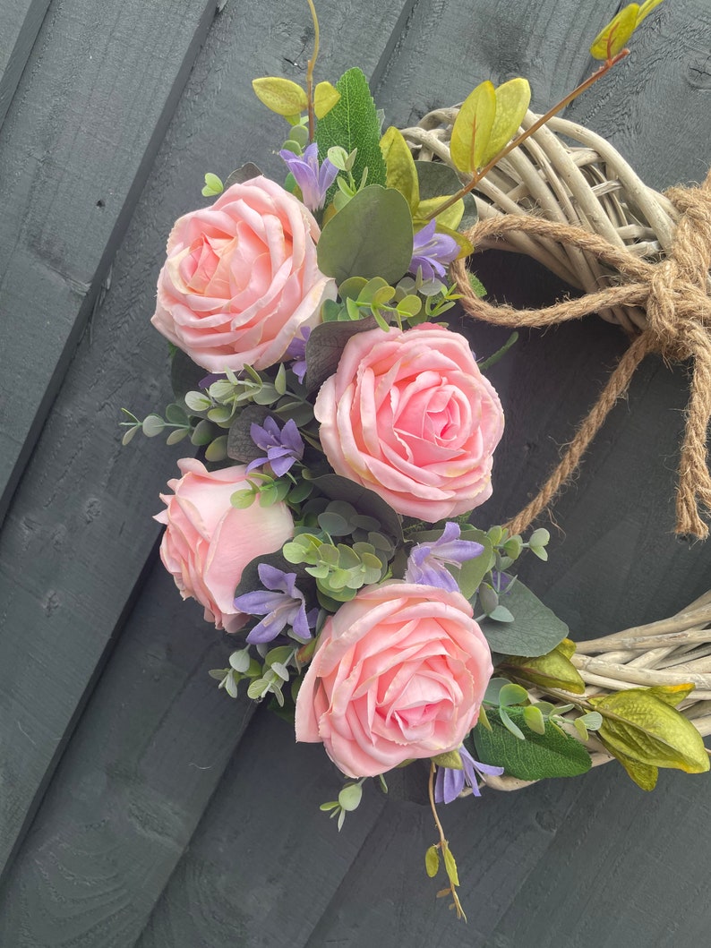 May include: A heart-shaped wicker wreath decorated with pink roses and purple flowers. The wreath is tied with a brown twine.