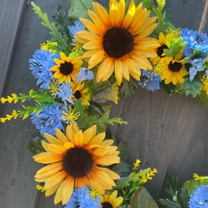 May include: A wreath made of artificial sunflowers and blue flowers. The wreath is made of green leaves and stems.