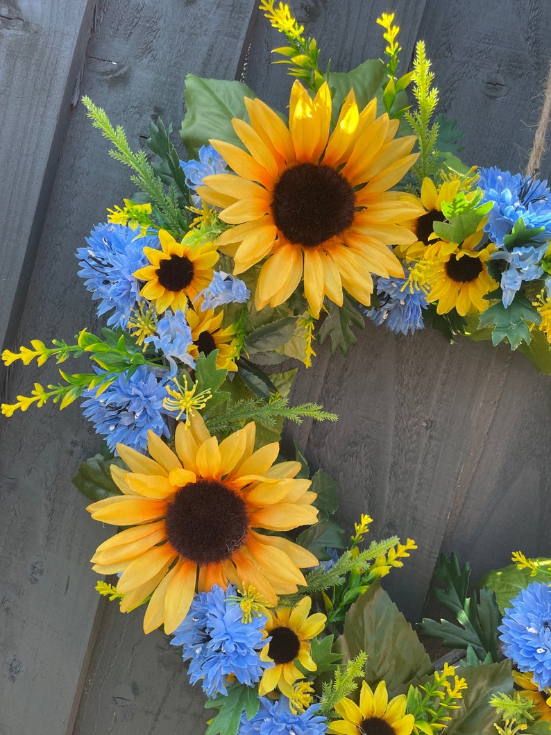 May include: A wreath made of artificial sunflowers and blue flowers. The wreath is made of green leaves and stems.