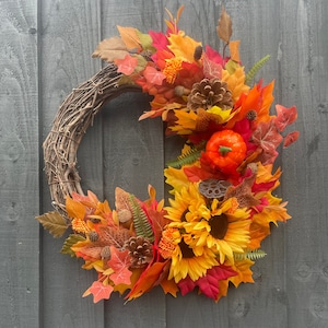 May include: A autumn-themed wreath with a brown twig base, decorated with faux leaves in shades of orange, red, and yellow. The wreath also features a small faux pumpkin, pinecones, and sunflowers.