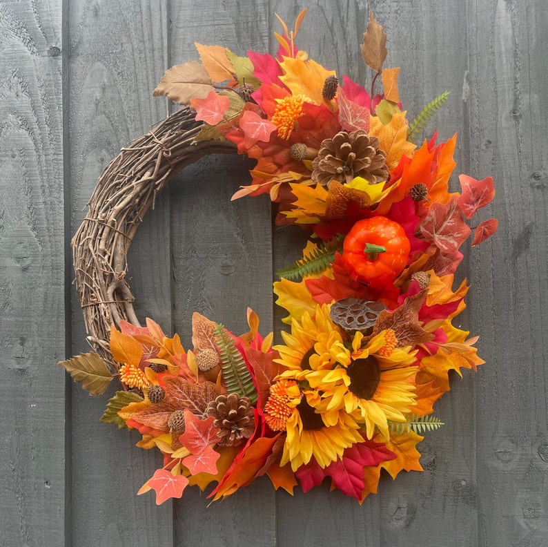 May include: A autumn-themed wreath with a brown twig base, decorated with faux leaves in shades of orange, red, and yellow. The wreath also features a small faux pumpkin, pinecones, and sunflowers.