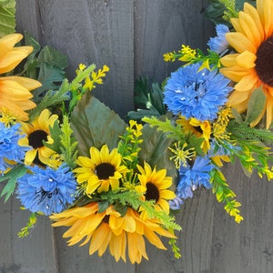 May include: A wreath made of yellow sunflowers and blue flowers with green leaves. The wreath is hanging on a grey wooden background.