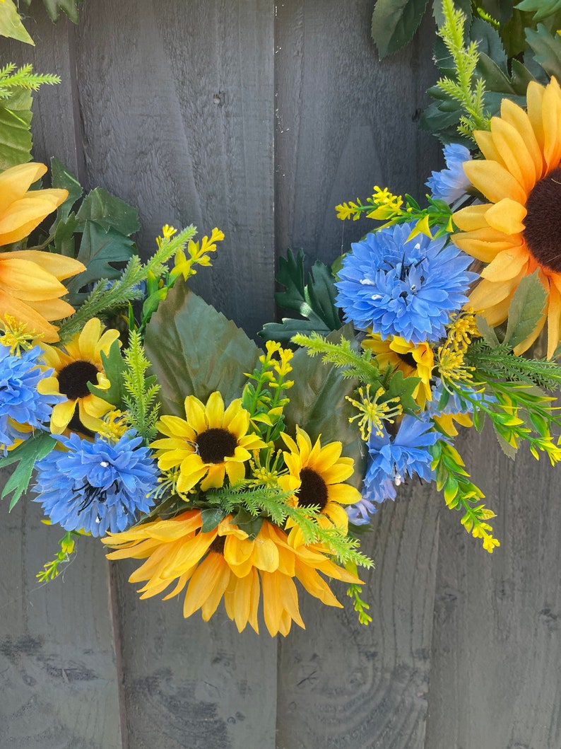 May include: A wreath made of yellow sunflowers and blue flowers with green leaves. The wreath is hanging on a grey wooden background.