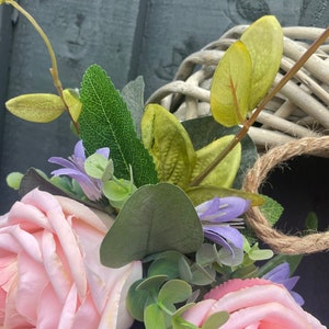 May include: A pink and white floral wreath with green leaves and a woven wicker base. The wreath is decorated with pink roses, purple flowers, and green eucalyptus leaves.