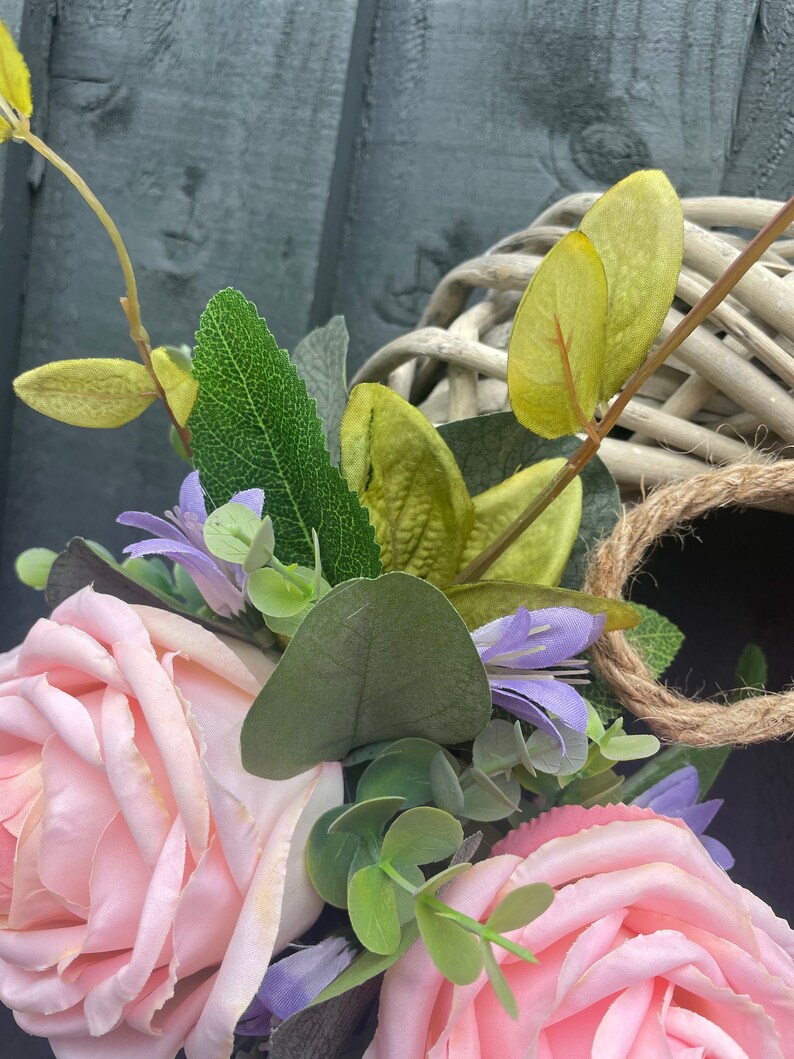 May include: A pink and white floral wreath with green leaves and a woven wicker base. The wreath is decorated with pink roses, purple flowers, and green eucalyptus leaves.